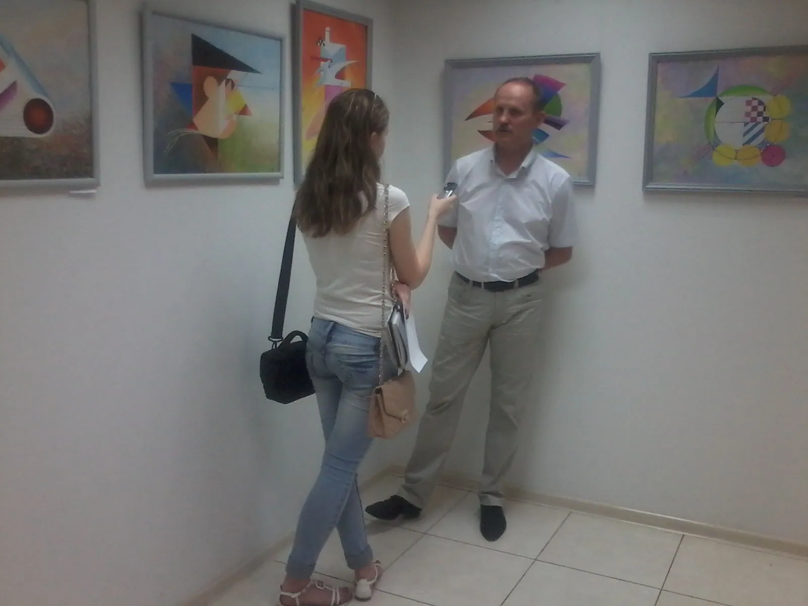 Oleksandr Zinovyev talking to a journalist in the gallery, his paintings on the wall behind him